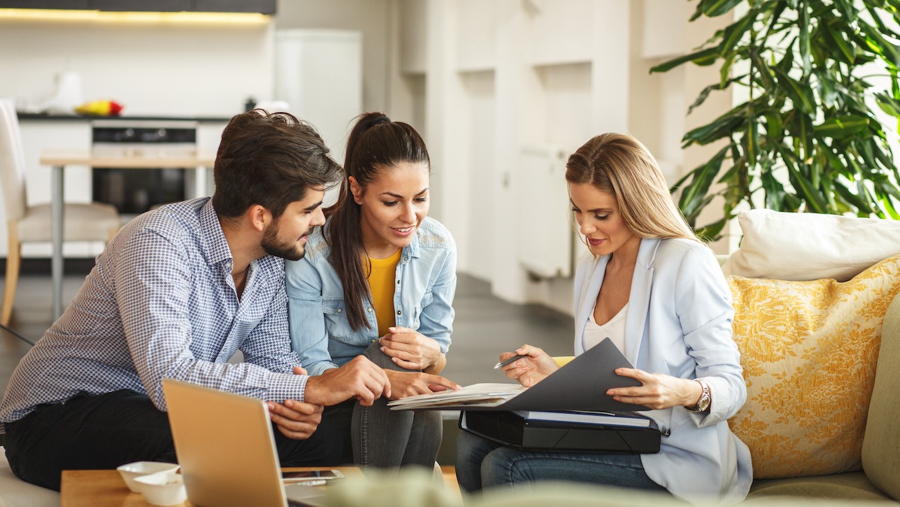 A wide-angle image of a confident real estate investor or small team reviewing floor plans or renovation plans on a table—with a laptop open showing rental data, and a property visible through the window or on-screen.